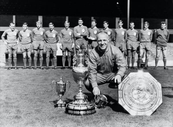 Liverpool manager Bill Shankly crouches by the trophies that his team won the previous season, including the League Championship trophy and the FA Charity Shield, as his players line up in the background: (lr) Ian St John, Ian Callaghan, Roger Hunt, Gordon Milne, Peter Thompson, Ron Yeats, Chris Lawler, Tommy Smith, Geoff Strong, Gerry Byrne, Willie Stevenson, Tommy Lawrence. 1966. (PA Photos/PA Archive/PA Images)